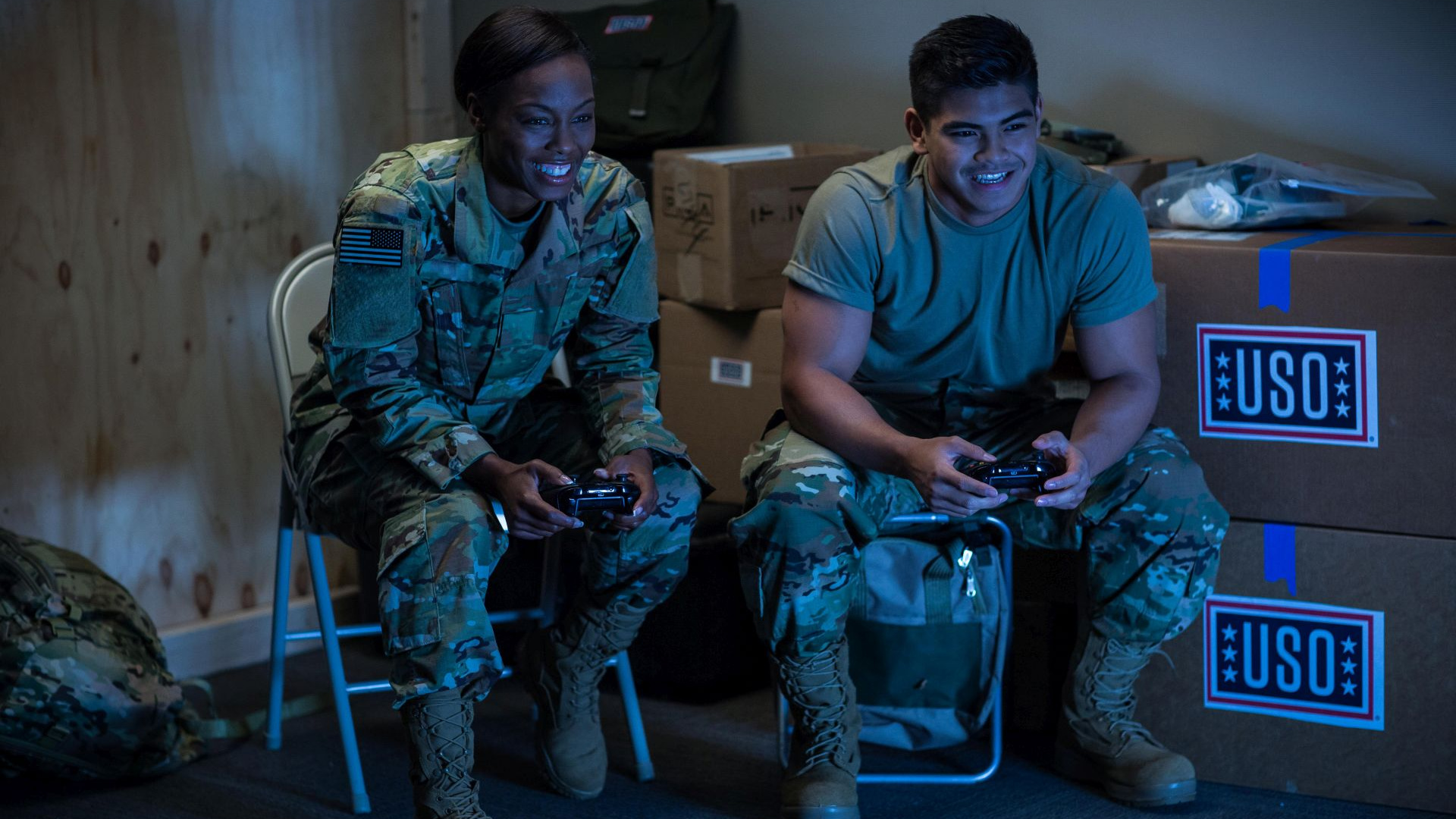 Two service members smiling and sitting in metal chairs playing a video game.