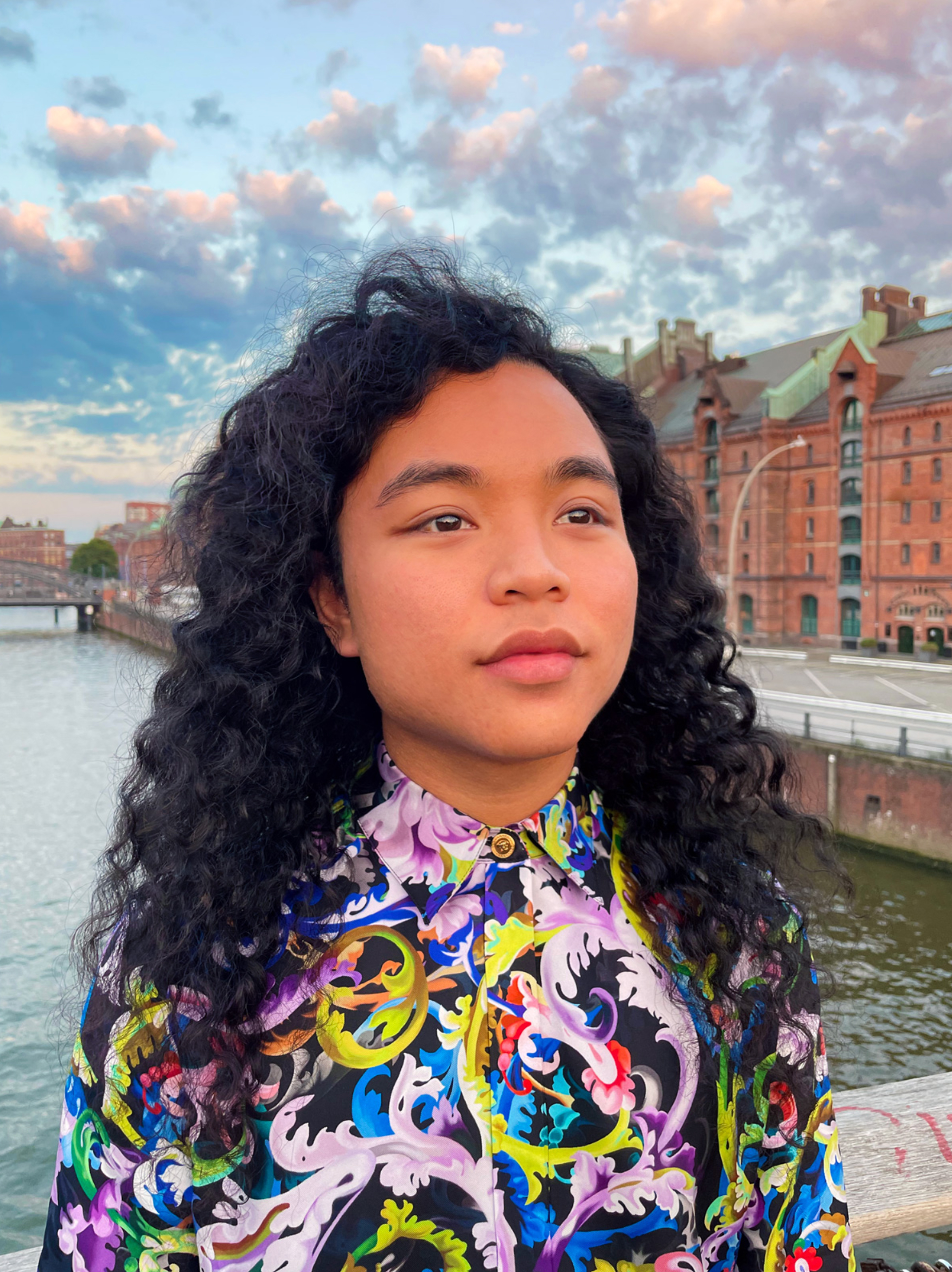 Portrait of artist Ari Liloan wearing a colorful patterned shirt near a waterside with brick buildings.