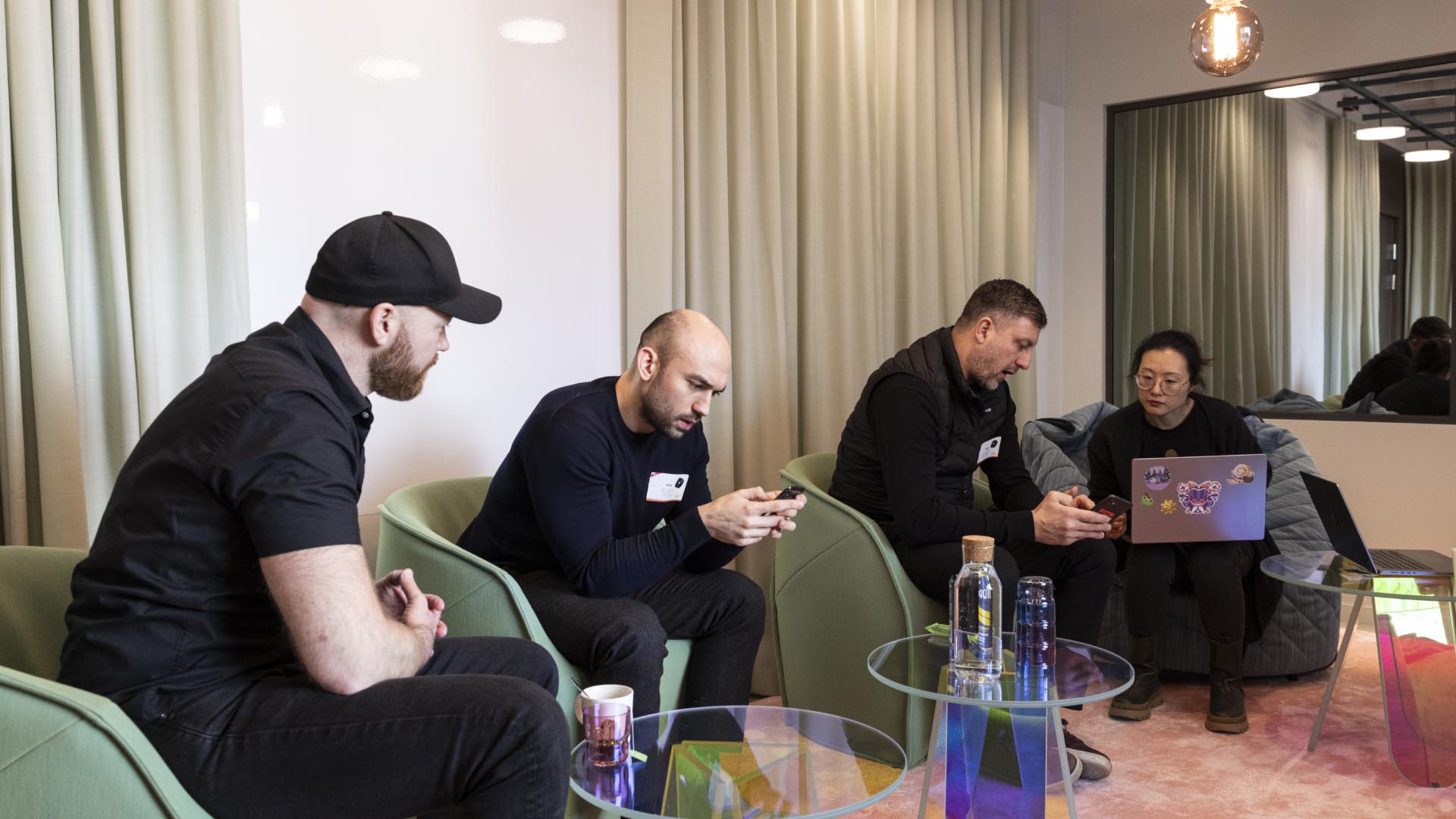 A group of four people huddling in a room, on their phones and computers.