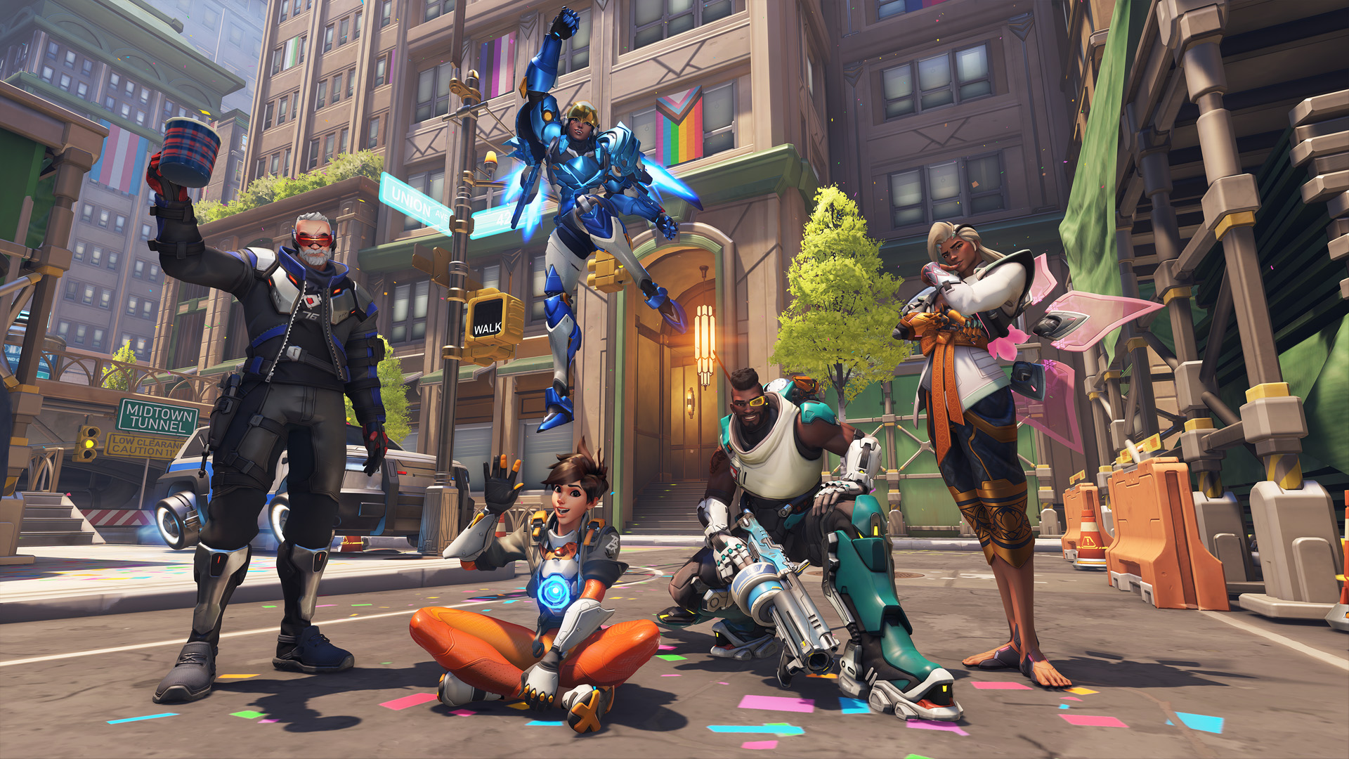Weitwinkelaufnahme der Karte Midtown von Overwatch 2, angepasst mit regenbogenfarbenem Thema und Pride-Flaggen. Von links nach rechts: Soldier 76 hebt einen Becher, Tracer winkt, während sie im Schneidersitz sitzt, Pharah feiert in der Luft, Baptiste hockt und Lifeweaver posiert für das Foto.