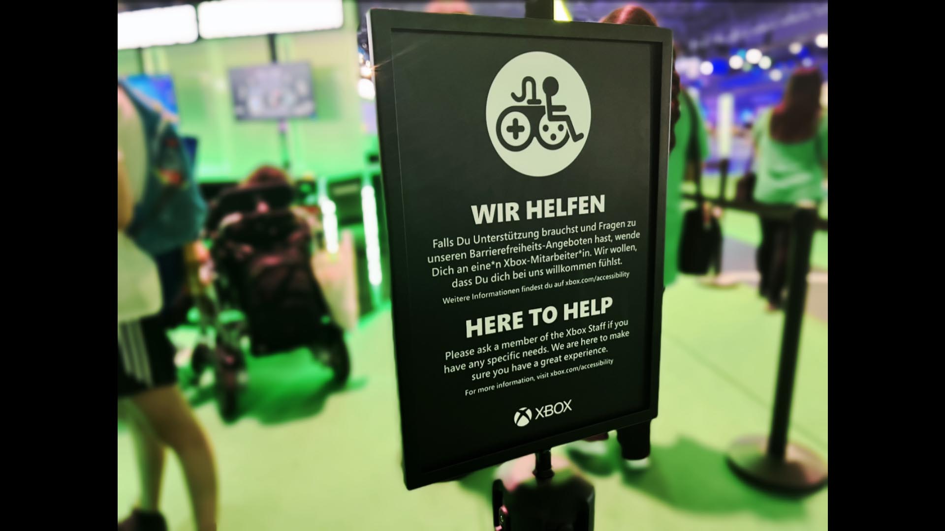 Ein schwarzes Schild mit einem weißen Logo für barrierefreies Spielen steht vor den Demostationen auf einer Spielemesse. Der Text lautet auf Englisch und Deutsch: "Hier wird dir geholfen. Bitte frage ein Mitglied des Xbox Teams, wenn Du besondere Bedürfnisse hast. Wir sind dafür da, dass Du ein tolles Erlebnis hast. Weitere Informationen findest Du unter xbox.com/accessibility. Xbox."